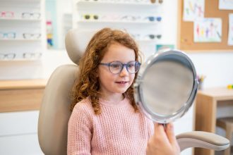 Petite fille essayant des lunettes rondes bleues chez un opticien à Brest, assise dans un fauteuil moderne avec un miroir devant elle